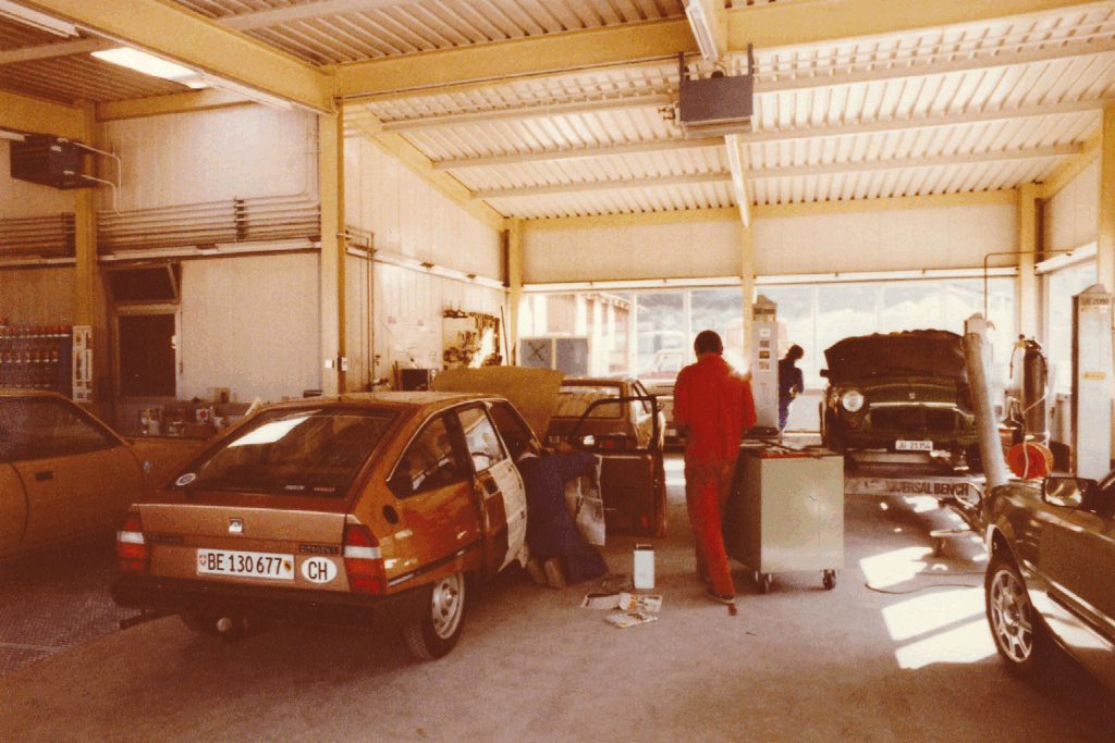 Photo d’archive de l’atelier de la Carrosserie du Relais SA à Sonceboz-Sombeval dans les années 1980, montrant les mécaniciens et carrossiers au travail sur plusieurs véhicules.